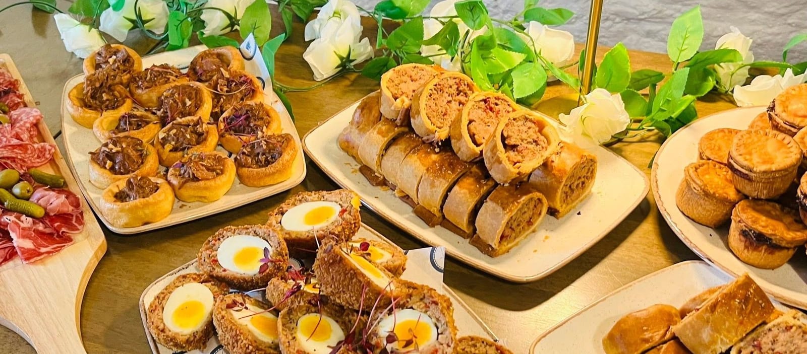 A party buffet on a table: Yorkshire pudding bites, sausage rolls, scotch eggs and pies, with flowers and greenery
