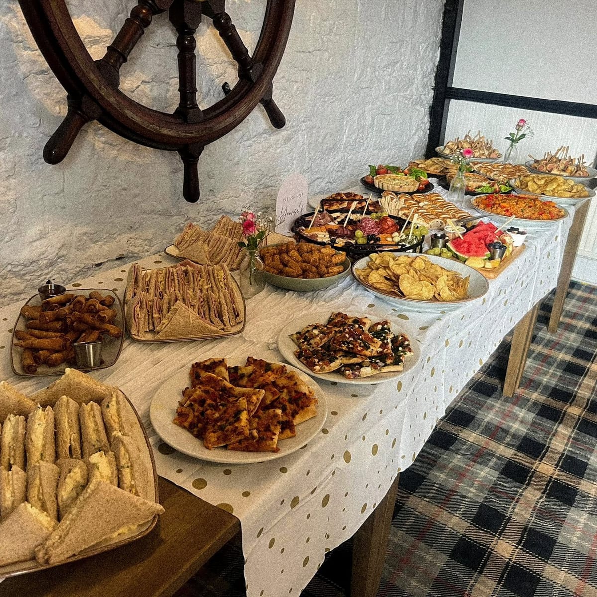 A long buffet table of sandwiches, hot dishes and platters laid out beneath a ship’s wheel on the wall