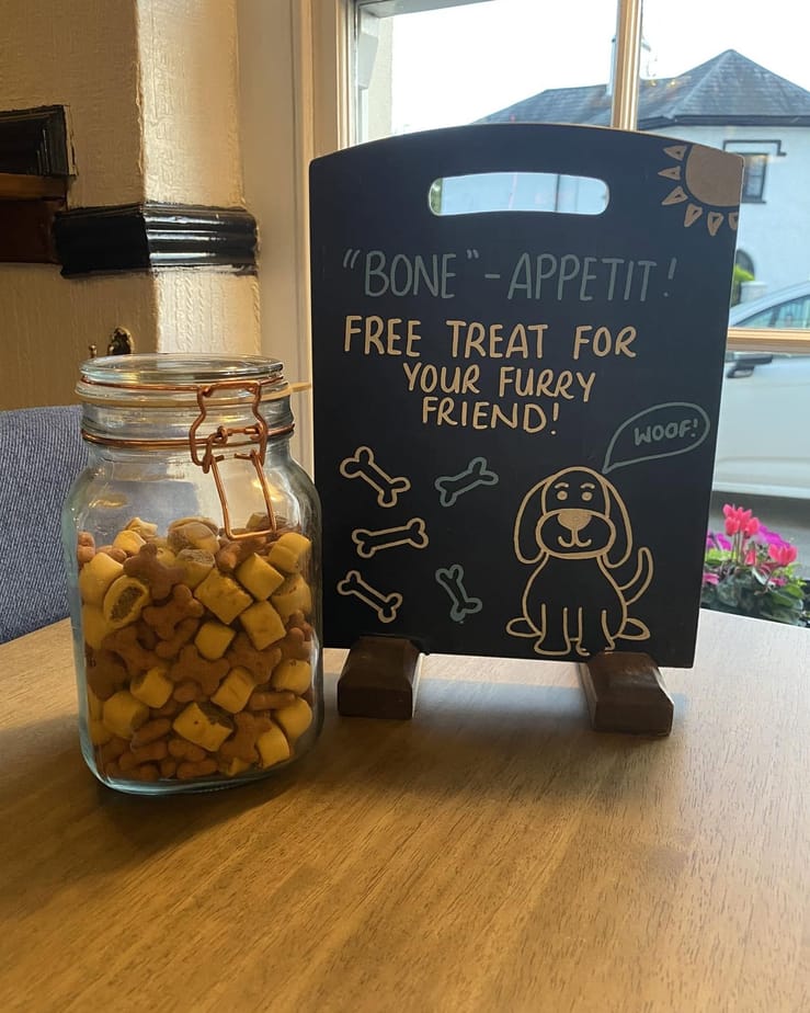 A jar of dog treats beside a chalkboard sign that reads “Bone-appetit! Free treat for your furry friend!”