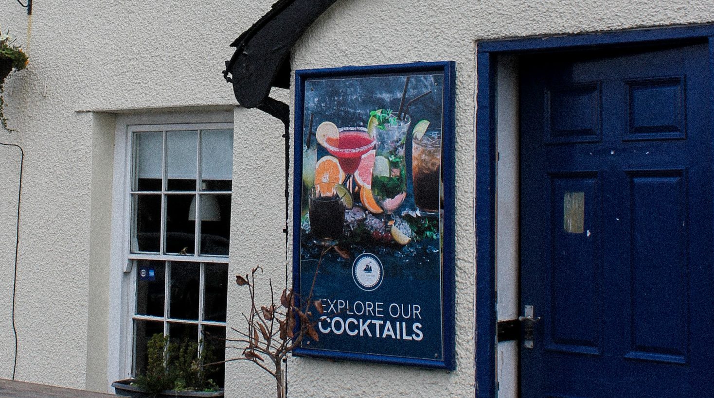The front of The Ship Inn: a sash window, a cocktails poster and the blue front door