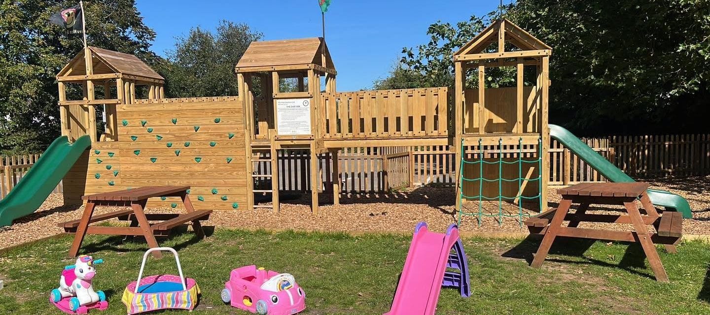 The wooden climbing frame with towers, a climbing wall and slides in the beer garden, with picnic tables and children’s toys on the lawn