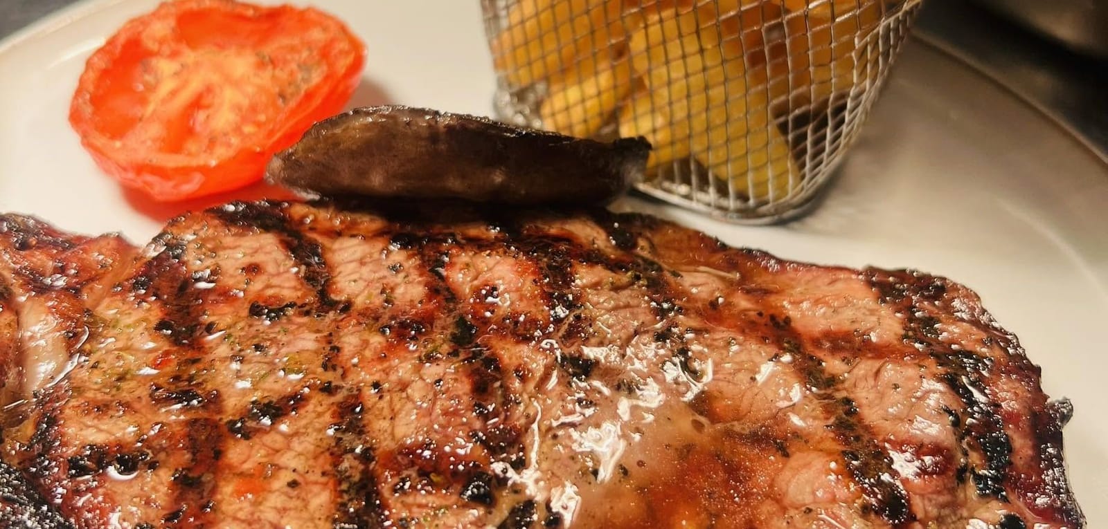 A grilled steak with a grilled tomato and mushroom, and a wire basket of chips
