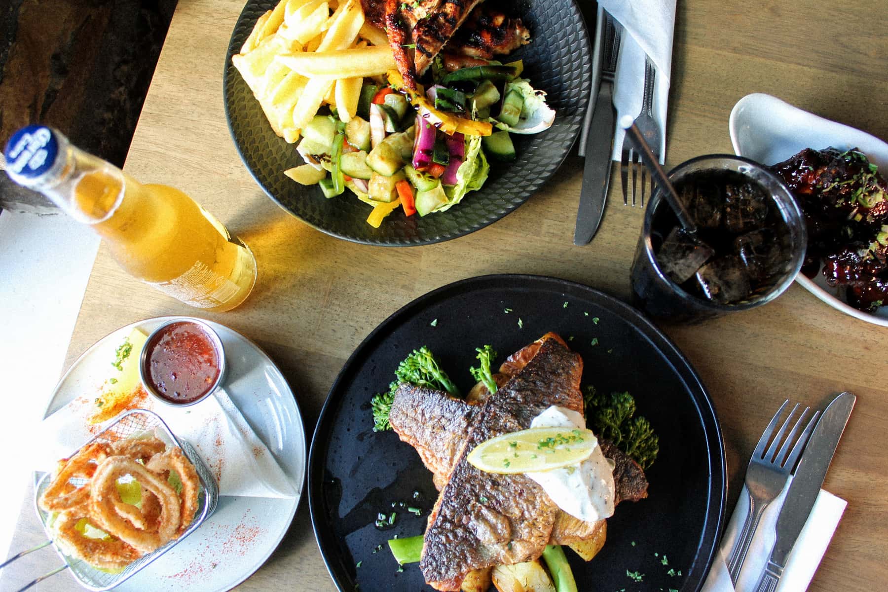 A table of dishes seen from above: pan-fried fish on a black plate, chicken with chips and salad, onion rings, glazed ribs and a bottle of beer